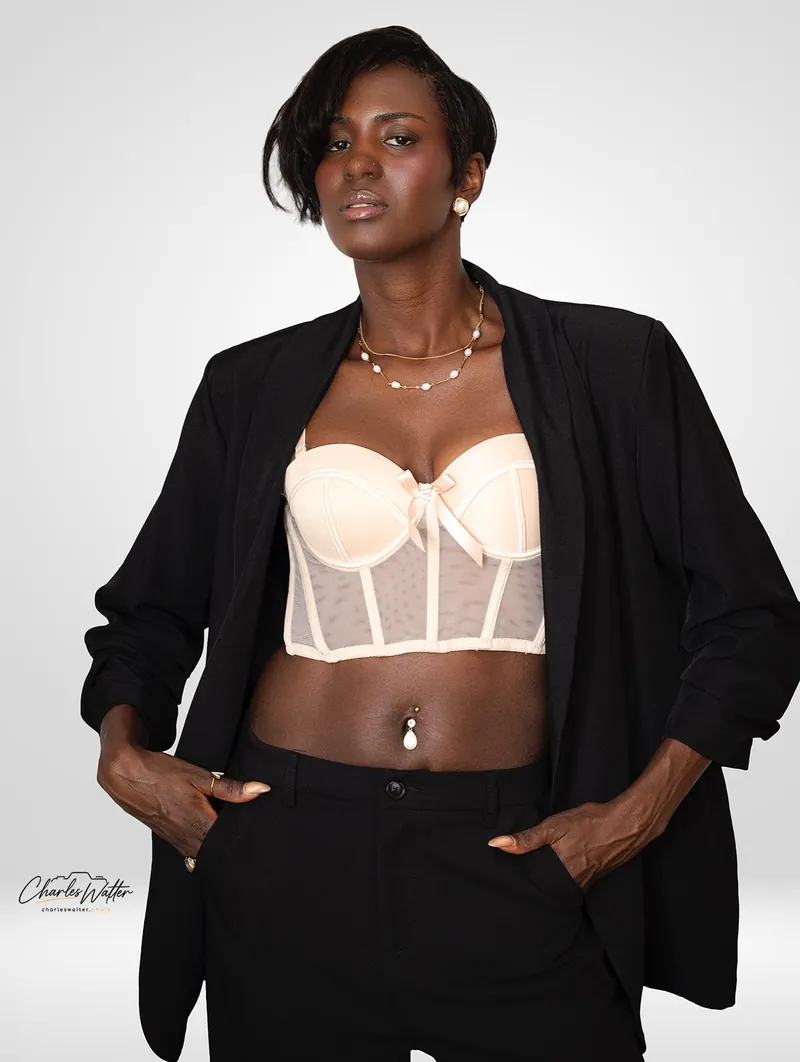 Retrato de estúdio em fundo claro: mulher de blazer preto aberto sobre corselete champagne, mãos nos bolsos.